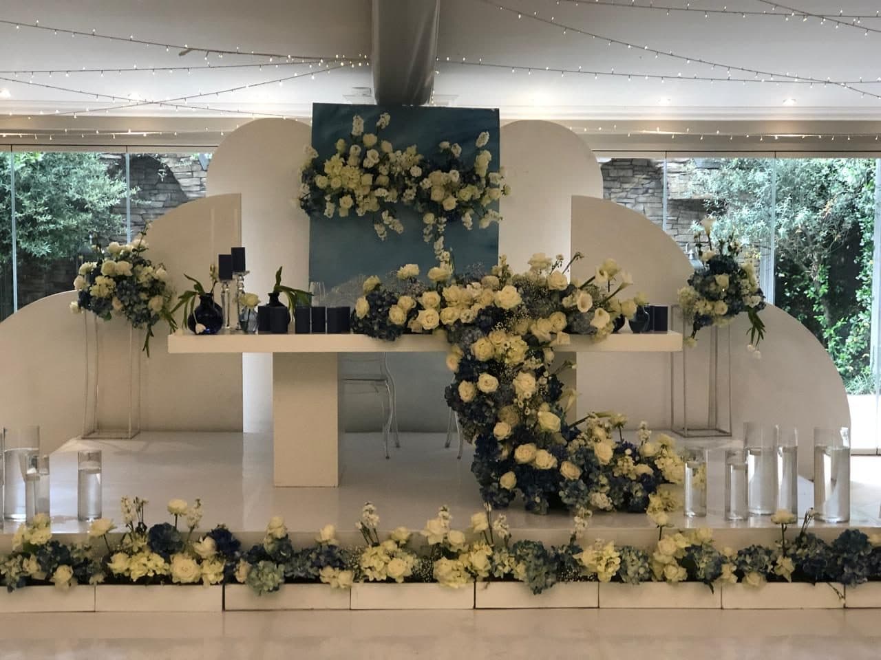 Arched bridal table backdrop banked with white roses, hydrangeas and navy vases