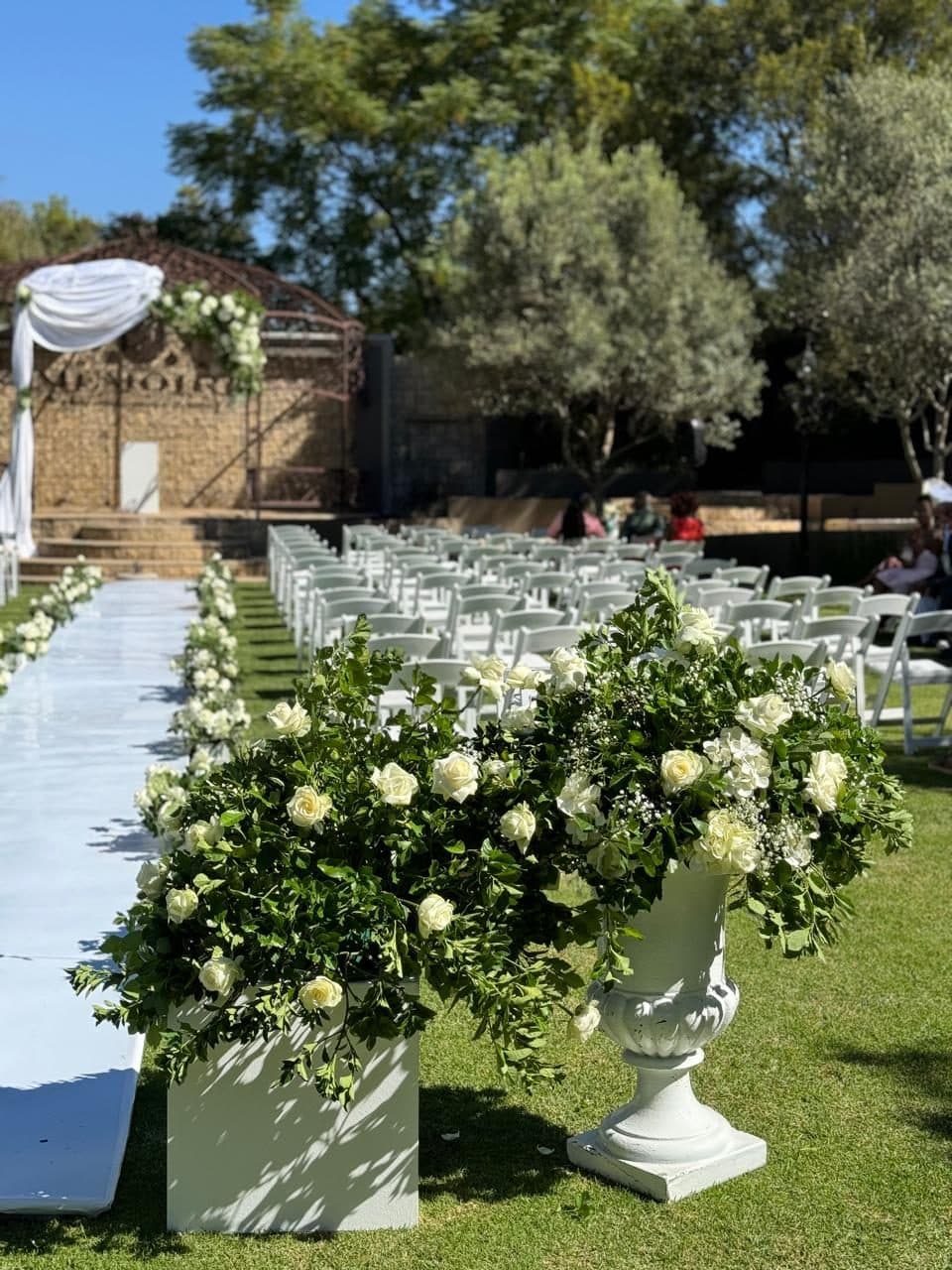 Outdoor ceremony aisle lined with white roses and greenery in stone urns