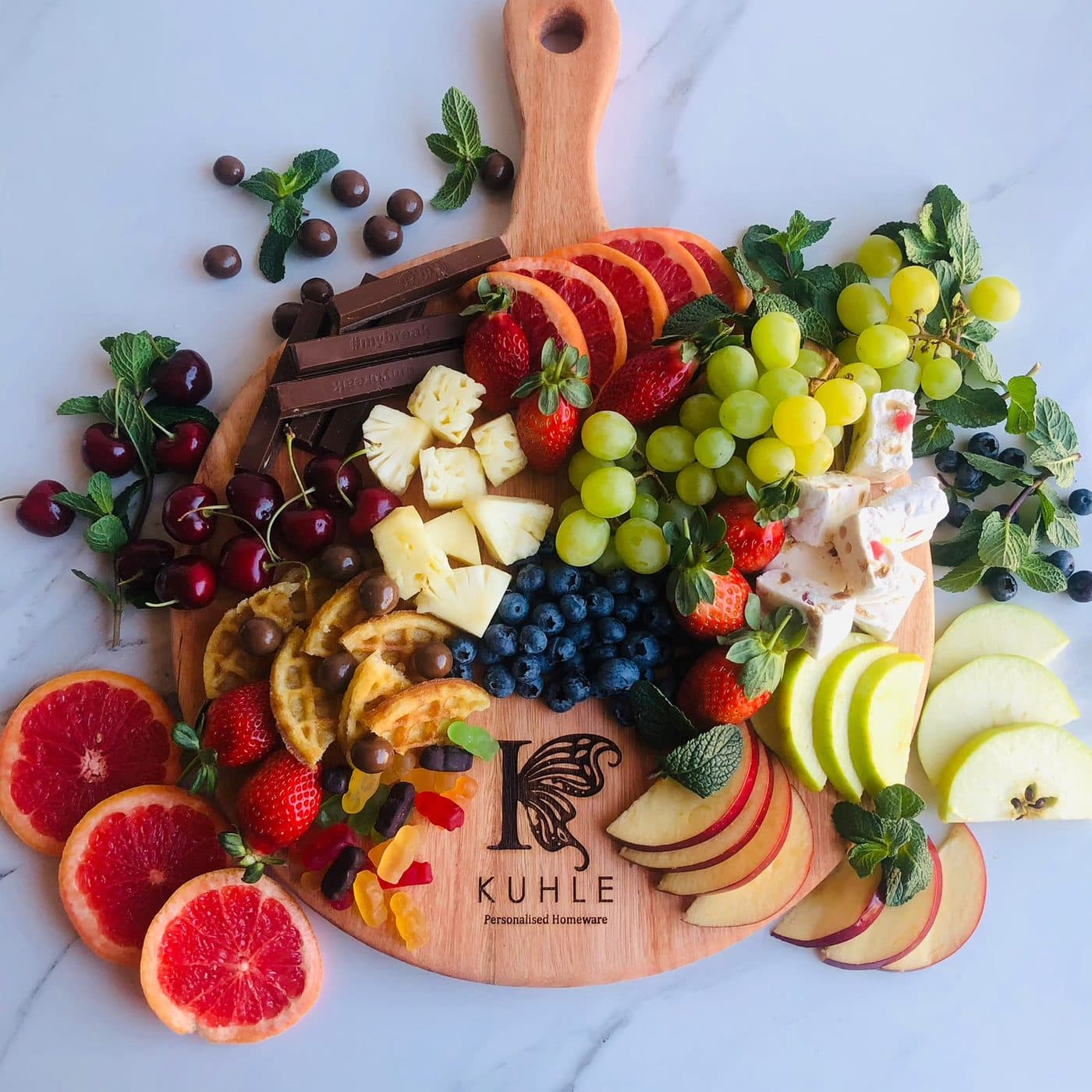 A round engraved Kuhle paddle board styled as a grazing platter with fruit, chocolate and nougat